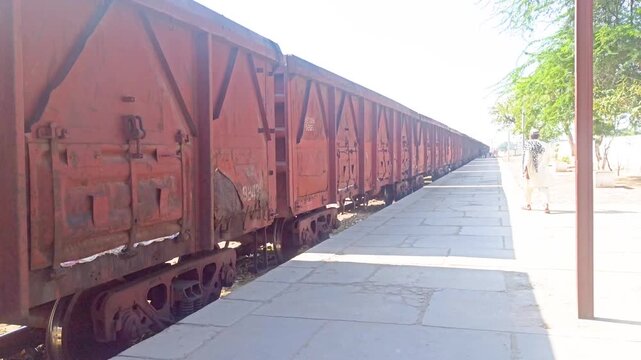 Long cargo freight train standing and passing by at a rural railway station platform in Pakistan while a local man in traditional shalwar kameez walks along the platform on a sunny day