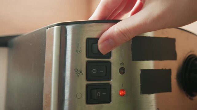 Close view of hand pressing top rectangular button on brushed stainless steel toaster control panel, with index finger, steady thumb grip, glowing red indicator light, and compact kitchen appliance