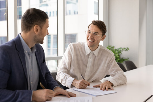 Happy businessmen signing contract at desk celebrating good deal satisfied with reaching profitable agreement enjoying successful partnership acknowledging mutual benefit positive negotiations results