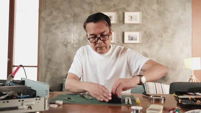 Mature Asian man voltage test at disassembling circuit board with multimeter tool, focusing on precision checks for home electrical appliance DIY fix, electronics component hobby, handyman workshop.