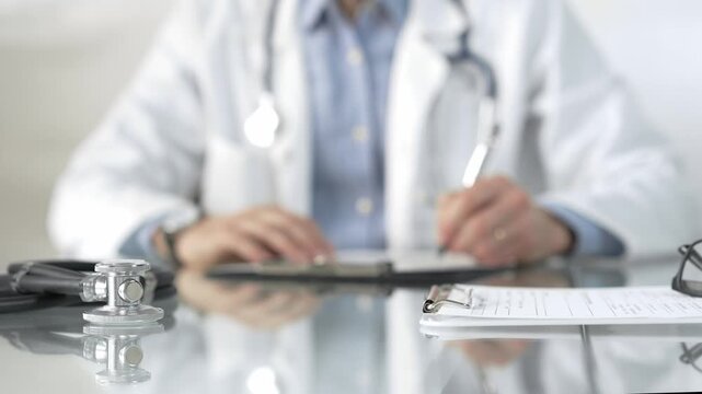 Stethoscope and medication history records form are on a glass table before a male doctor working with laptop in clinical office background. Medicine concept