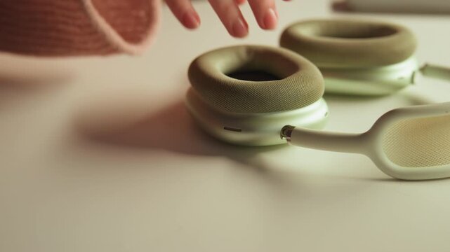 Close-up scene shows hand lifting retro wired earbud from pale tabletop, exposing worn grille, foam pad, textured rim, muted finish, soft shadow, and warm side light across vintage audio surface