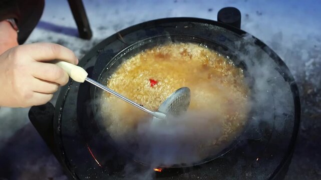 Traditional plov cooking outdoors in winter, with rice simmering in a black pan while a person stirs the hot dish over a wood stove surrounded by snow and open flames.