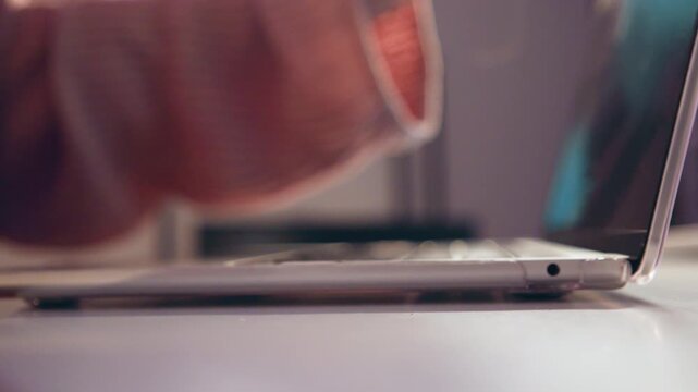 Low angle close-up of slim metallic smartphone on white tabletop beside laptop with lid rising behind, while sleeve movement suggests brief user interaction in soft indoor light and blurred background