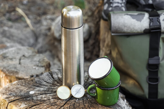 Adventure travel and outdoor exploration, stainless steel thermos with enamel mugs and compass on a tree stump for hiking and wilderness camping trip