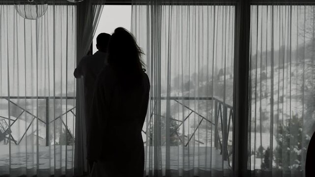 Woman approaches a man and hugs him by a large window overlooking snowy mountains. Silhouetted couple sharing a tender moment during a winter getaway.
