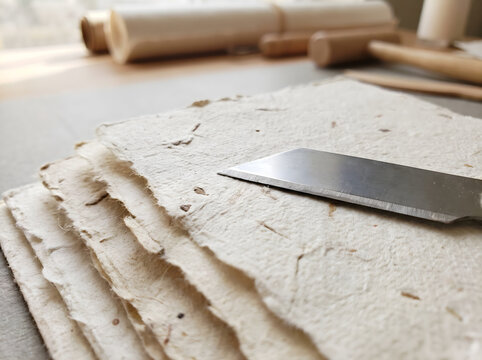 A sharp craft knife or exacto blade resting on a stack of handmade deckle edge paper in a bookbinding studio