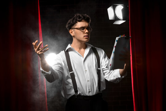 Acting audition and theater performance, young male actor rehearsing lines with script on stage under spotlight with dramatic smoke and red curtains