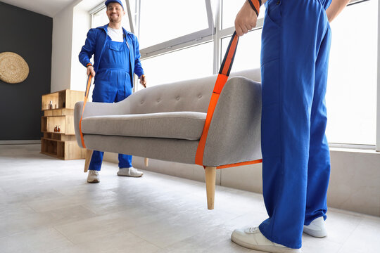 Moving service and relocation, professional movers carrying a sofa with lifting straps in a new home interior for residential furniture delivery