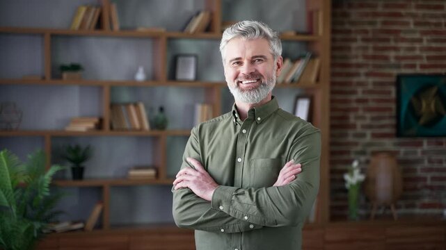 Portrait of bearded man with gray hair smiling confidently while standing with crossed arms in home office. Happy male showing positivity and success, looking at camera with calm, friendly expression
