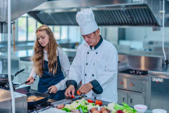 Senior Asian chef teaching teen culinary intern to fry food on stove in a professional kitchen