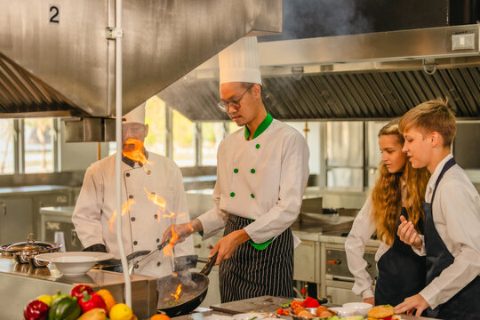 Asian chef cooking with flaming pan as teen culinary interns watch in a professional kitchen