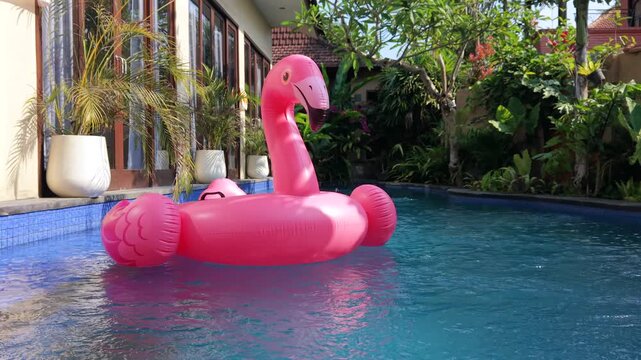 Pink flamingo inflatable floating in a tropical swimming pool surrounded by lush greenery. Colorful summer vacation, resort, leisure and tropical lifestyle concept.