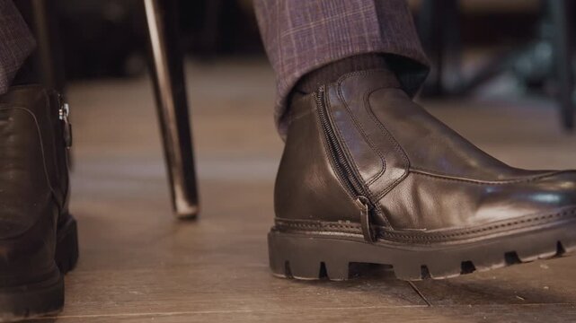 Close up of man hand adjusting zipper on his black leather ankle boot while seated. focus remains solely on foot and shoe highlighting details like stitching texture detail and side zipper
