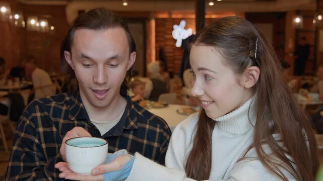Close up of man and young woman in restaurant setting with girl offering him ceramic cup. scene shows warm indoor lighting as they interact casually among other diners at tables. Background