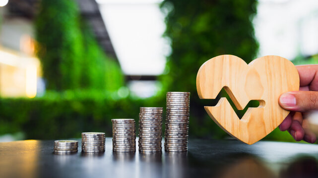Health insurance and medical savings concept, hand holding wooden heart with pulse rhythm beside ascending stacks of coins on table, symbolizing healthcare funds, wellness, and treatment.