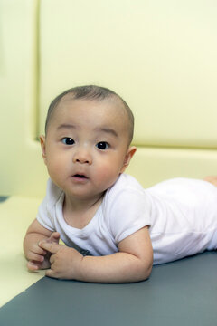 Infant tummy time and newborn milestones concept, cute Asian baby boy in white onesie lying on playroom floor mat looking at camera, representing early childhood care, and pediatric health.