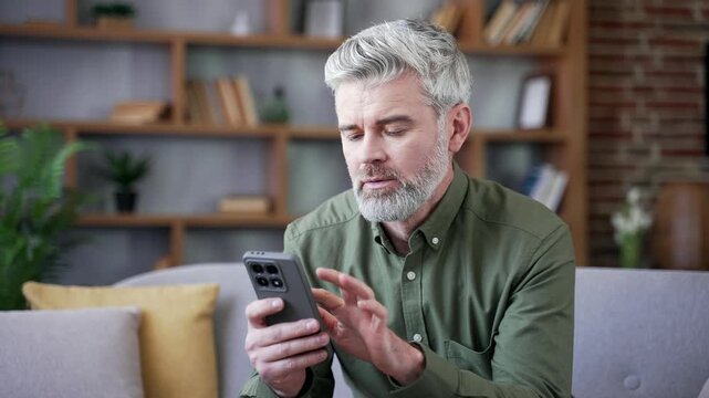 Close-up of thoughtful mature bearded man with gray hair using smartphone at home. Serious male looking at screen reading important news or message, reflecting quietly in cozy living room atmosphere