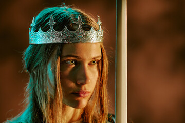 Obraz premium Young adult Caucasian woman wearing silver crown holding sword, looking away with serious expression. Concept representing power, leadership, suitable for fantasy gaming and storytelling