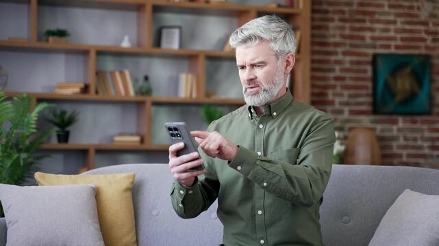 Frustrated bearded man with gray hair sitting on sofa at home using smartphone. Senior male trying to make online payment but facing technical issue, looking confused, disappointed at screen indoors