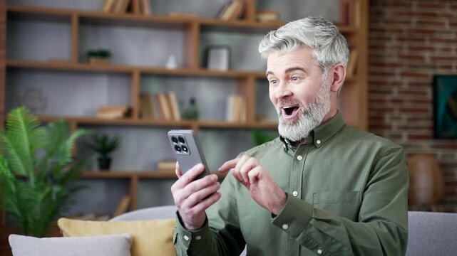 Close-up of bearded man with gray hair smiling after reading great news on smartphone at home. Excited male feeling happy and surprised, enjoying positive emotions and success in cozy living room