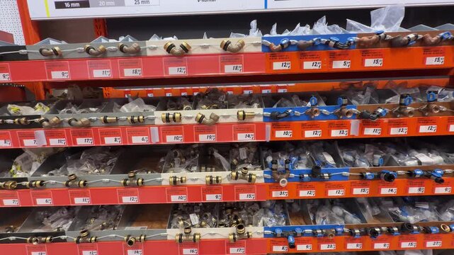 Panning shot across hardware store shelves displaying assorted plumbing fittings, including brass compression fittings, elbows, couplings, adapters, connectors and flexible hoses with retail price lab