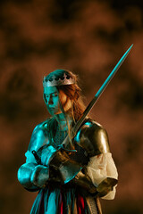 Obraz premium Vertical image of Caucasian young adult woman wearing medieval knight armor with crown holding sword looking down. Female warrior concept useful for fantasy game promotion, vertical