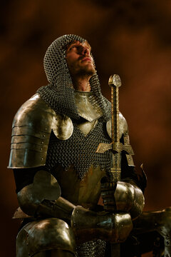 Vertical image of Caucasian young adult man in metal armor holding sword while looking upward, representing honor and bravery for fantasy game marketing