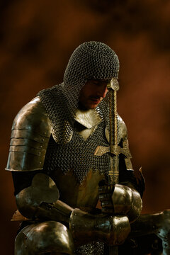 Vertical shot of Caucasian young adult man wearing medieval armor leaning on sword while praying. Concept image suitable for historical gaming advertising or fantasy book covers