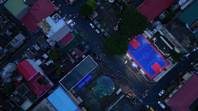 Cinematic aerial footage of a brightly illuminated basketball court during a community event in Buhi, Camarines Sur, Philippines. Captured from a distant perspective at blue hour, the sports court app
