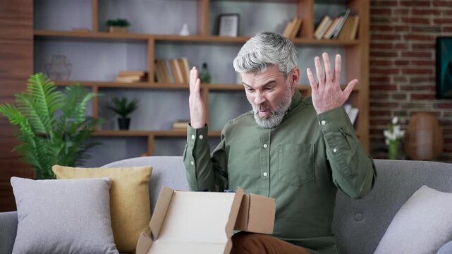 Man with gray hair sitting on sofa at home looking disappointed after opening a package. Unhappy male customer dissatisfied with online purchase, expressing frustration and discontent in living room