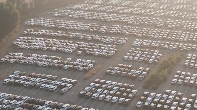 Car storage yard contains hundreds of parked vehicles arranged in dense parallel rows across a large gravel lot with scattered vegetation, aerial drone slide