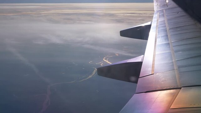 Commercial passenger airplane wing flying high above a winding river landscape and light clouds during a sunset flight. Beautiful scenic natural background template for global travel, international. 