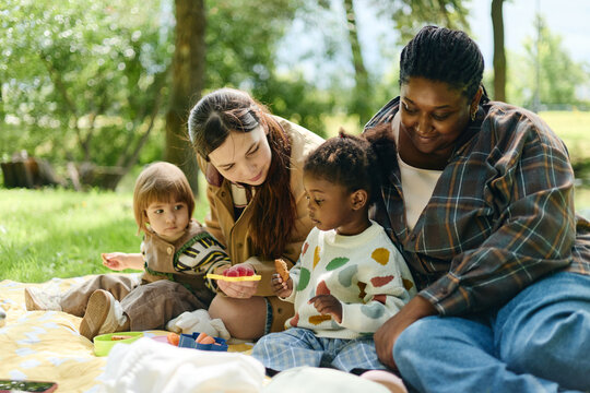 Diverse African American and Caucasian mothers enjoying picnic with toddler daughters in park. Concept representing multicultural parenting, family friendship, modern lifestyle marketing