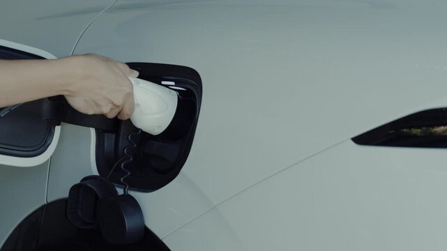 Close up shot of hand plugging power charger into white electric vehicle charging port at home.