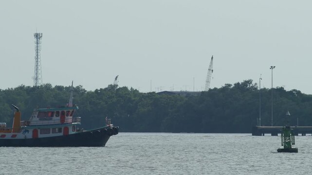 Colorful tugboat towing a small fishing boat with rope across calm blue sea, green navigation buoy and seagull on horizon