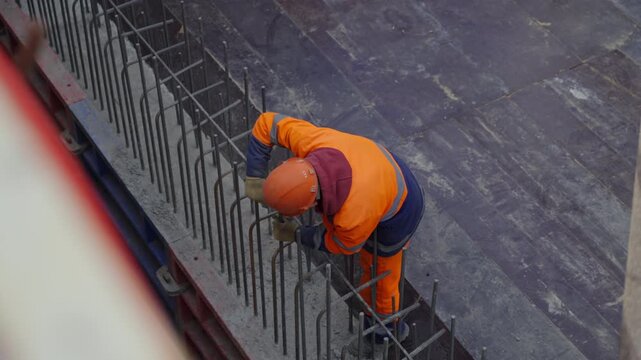 Construction worker ties metal rebar together on tall concrete structure. Individual wears orange high-visibility uniform and helmet