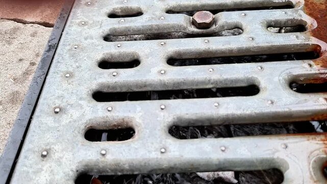 Weathered metal drainage grate with elongated openings, rust, dirt, and a small stone resting on the surface, installed along an urban pavement drainage system