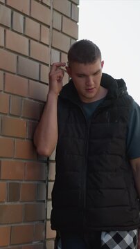 Close up of young man standing near brick wall holding and using vape device. Man wears black puffer vest over t shirt with checkered shorts visible. Soft natural light illuminates outdoor