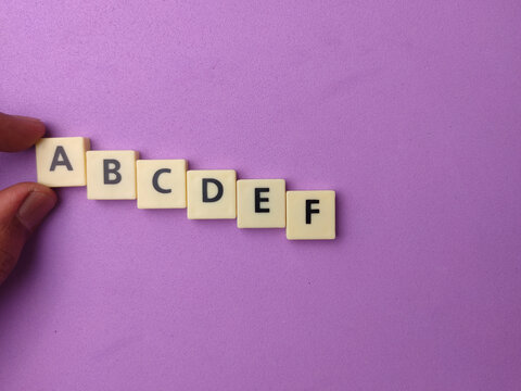 A hand holds letter tiles spelling the alphabet sequence A to F on a purple background