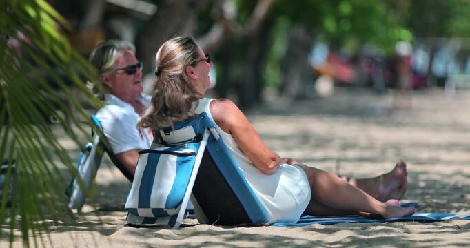 Mature couple relaxing in beach chairs under palm trees on a sunny tropical shore, enjoying a peaceful resort getaway together, overlooking the calm blue sea. Travel vacation to Samui Island, Thailand