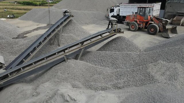 Heavy Machinery Orange Wheel Loader Near a Rock Crusher Machine with Conveyor Belt System and Aggregate Stockpiles Viewed from Above in Banyumas, Central Java, Indonesia.