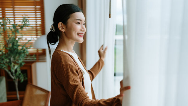 Happy Asian woman standing by the window and looking outside the living room enjoying the bright sunshine
