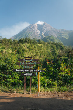 Wooden directional signpost along a trail with Mount Merapi volcano and white smoke in the background under a blue sky