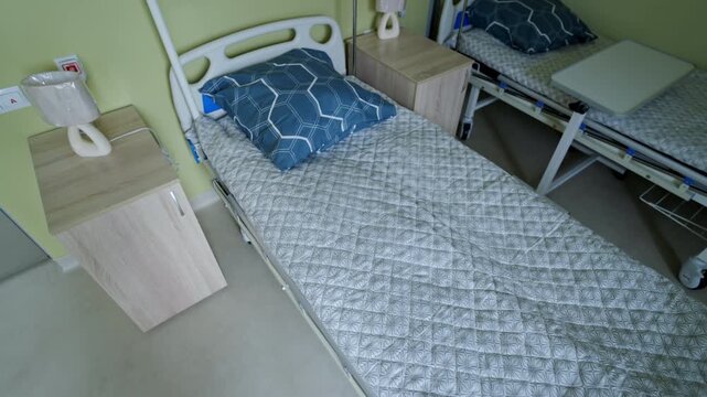 Medical bed features patterned blue pillow and quilted grey mattress cover. Wooden nightstand holds modern white table lamp nearby