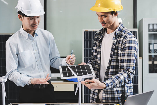 Two Asian engineers wearing hardhats and safety vests discussing solar panel installation and renewable energy project in the office.
