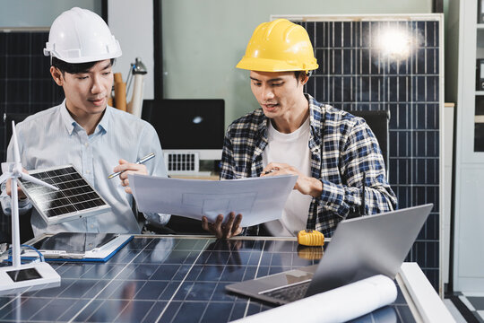 Two Asian engineers wearing hardhats and safety vests discussing solar panel installation and renewable energy project in the office.