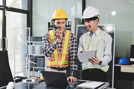 Two Asian engineers wearing hardhats and safety vests discussing solar panel installation and renewable energy project in the office.