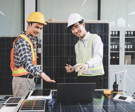 Two Asian engineers wearing hardhats and safety vests discussing solar panel installation and renewable energy project in the office.