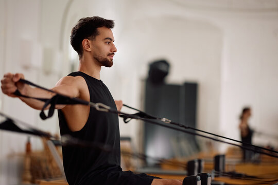 Male athlete exercising with resistance straps while working out or reformer machine in Pilates studio.